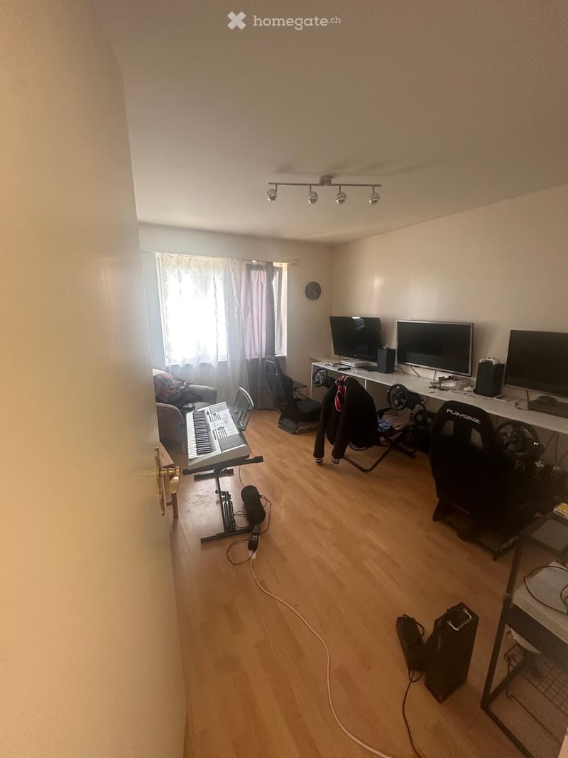 Empty room with wooden floor, white walls, a piano, two computer monitors, a chair, and a desk.