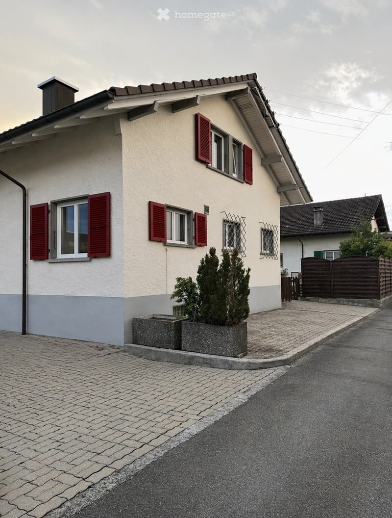 2 story house, white and gray exterior, red shutters, brick driveway, small garden with plants