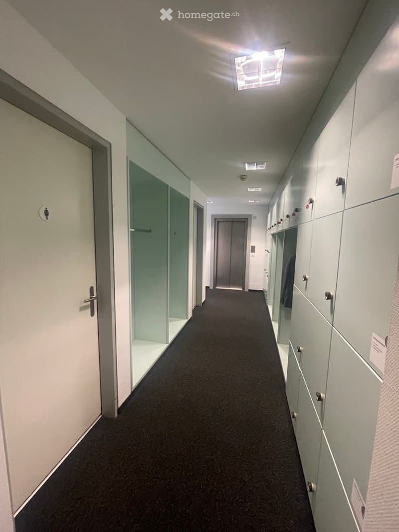 Empty hallway with white walls, black carpet, glass doors, lockers, and ceiling lights
