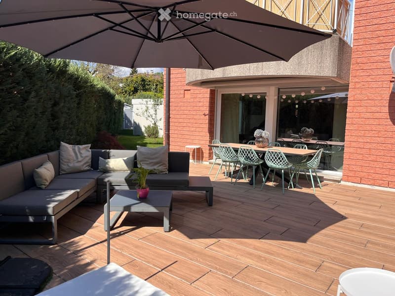 Wooden deck patio with grey sectional sofa, coffee table, dining table, and chairs, with a large umbrella providing shade. The patio is adjacent to a modern house with large glass doors.