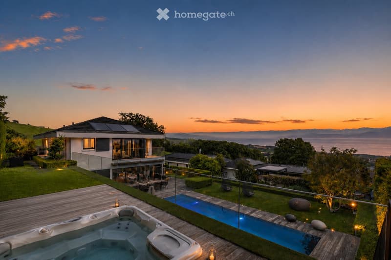 A modern house with a spacious outdoor area featuring a swimming pool, hot tub, wooden deck, and well-maintained lawn, overlooking a scenic lake and mountain view at sunset.