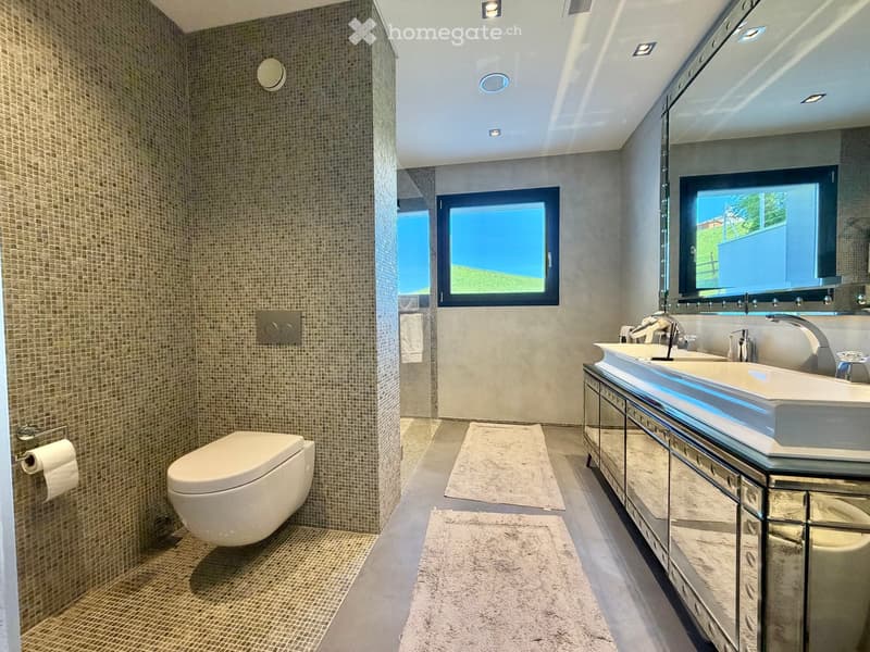 Modern bathroom with a toilet, double sink vanity, large mirror, and tiled walls. Features recessed lighting and a window with a view of greenery.