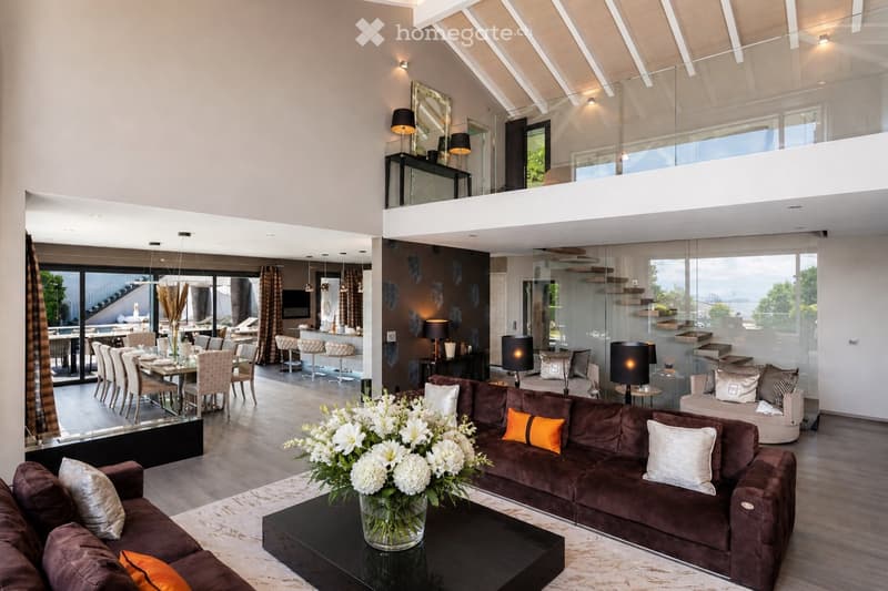Modern, open-concept living and dining area with high ceilings, large windows, and a balcony. Features a brown sectional sofa, coffee table with a flower arrangement, dining table with chairs, bar area, and decorative lighting.