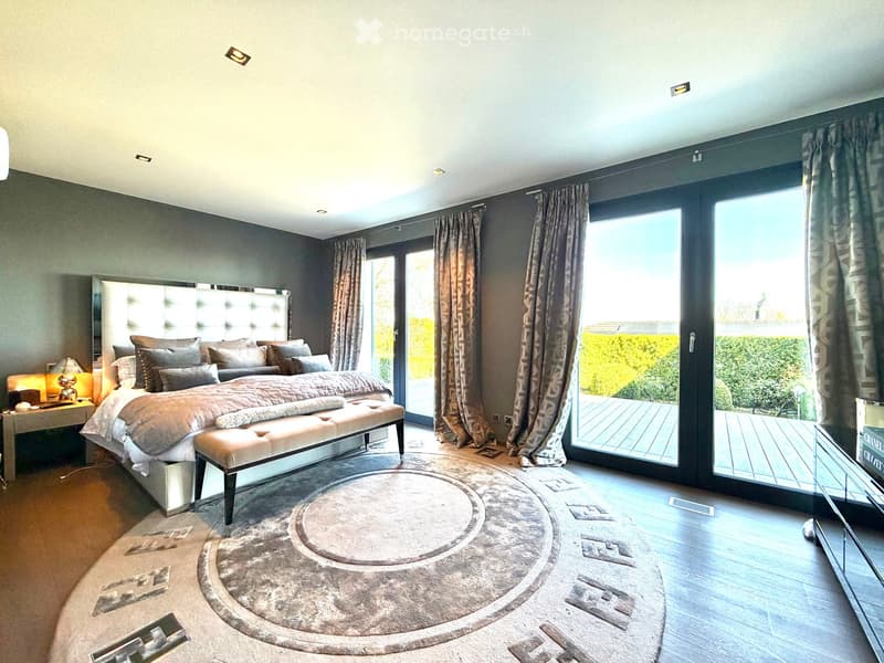 Modern bedroom with a large bed, bench, nightstand, lamp, and large windows with curtains leading to a balcony.