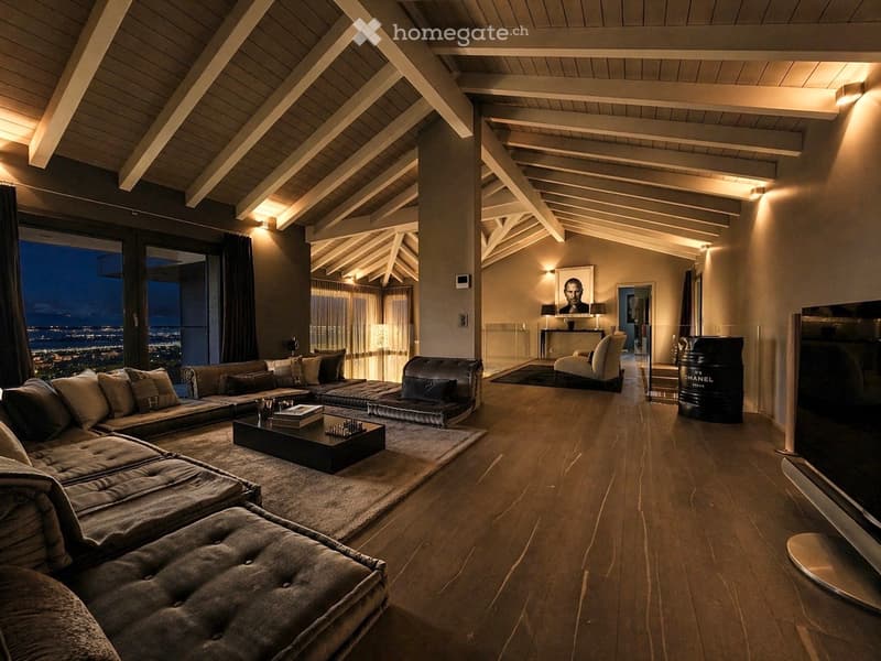 Modern living room with exposed wooden beams, large windows offering a city view, a sectional sofa, coffee table, ottoman, and a TV.