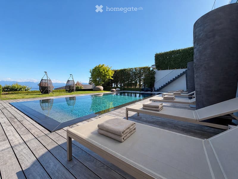 A modern outdoor area with a rectangular swimming pool, wooden decking, lounge chairs, and towels, surrounded by greenery and a clear blue sky.