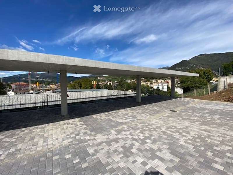 An outdoor parking structure is shown, with a concrete roof, a concrete deck with grey square paving stones, black railings, and a fence.