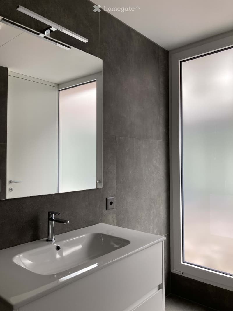 Modern bathroom with gray tiled walls, silver lighting, a white vanity, a glass mirror, a sleek silver faucet, and an open door with frosted glass.