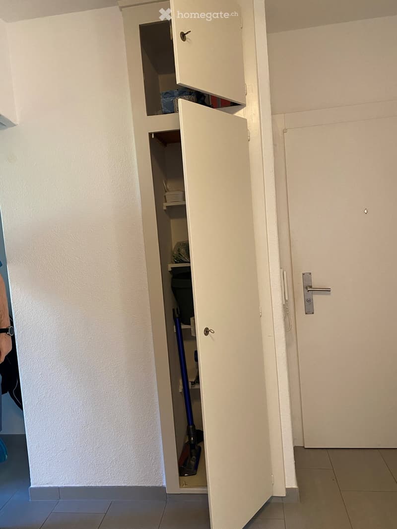 A tall white cabinet with two doors, one open, showing shelves and cleaning tools inside.