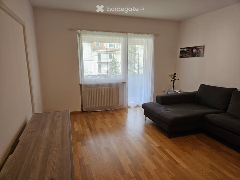 Living room with wooden floor, white walls, large window with white curtains, a black sofa, a wooden table, and a radiator.