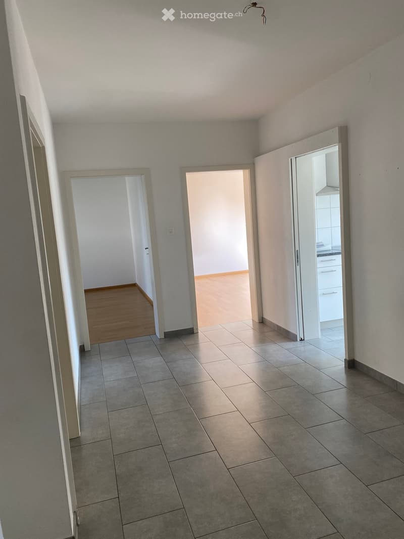 Empty hallway with tiled floor, white walls, doors leading to different rooms, wooden floor in one room, white kitchen cabinets