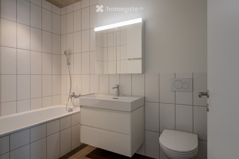 A modern and minimalistic bathroom with white tiles, a bathtub, sink with storage, a mirror, and a toilet.