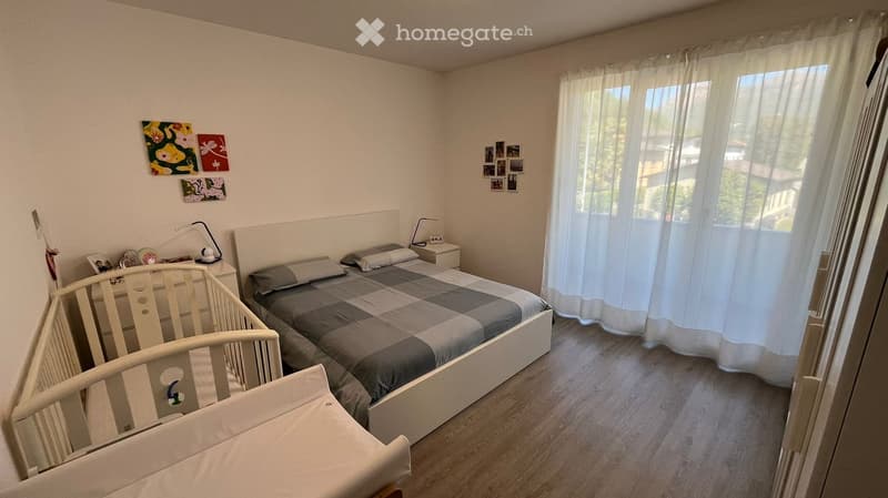 A bedroom with a bed, nightstands, lamps, and a crib. The walls are white, and there are picture frames and curtains on the window.