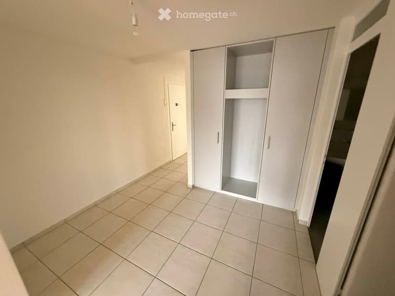 Empty room with white tiled floor, white walls, large white wardrobe, and a door leading to another room.