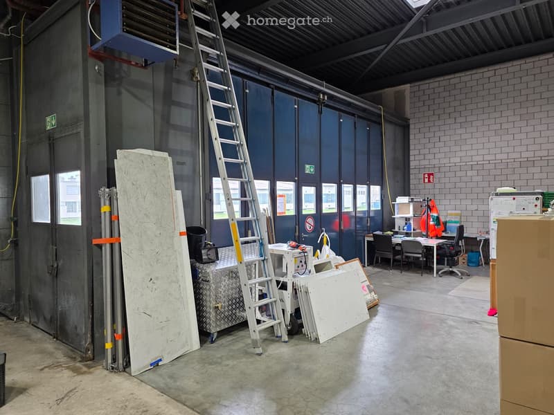 Industrial warehouse with large doors, metal storage boxes, ladders, and office furniture.
