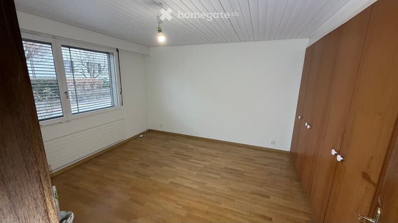 Large room, wooden floor, white walls, ceiling light, window with blinds, wooden cabinets.