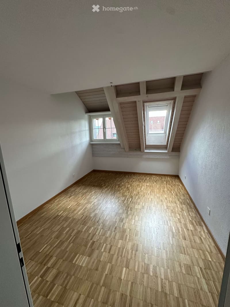 Empty room with wooden flooring, white walls, and two attic windows. No furniture or equipment.