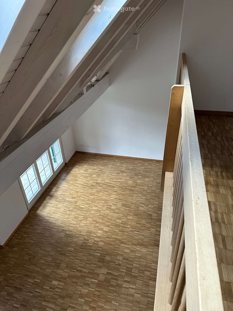 Empty room with wooden parquet flooring, white walls, wooden ceiling beams, and a window.