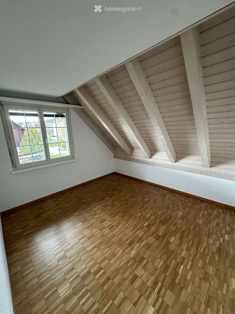 An empty attic room with wooden flooring and white walls. There is a window with white frames and a wooden ceiling with beams.