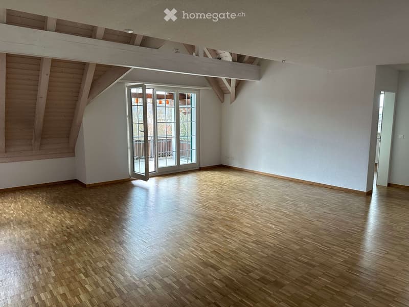 Empty room with wooden parquet flooring, white walls, wooden beams on the ceiling, and a glass door leading to a balcony.