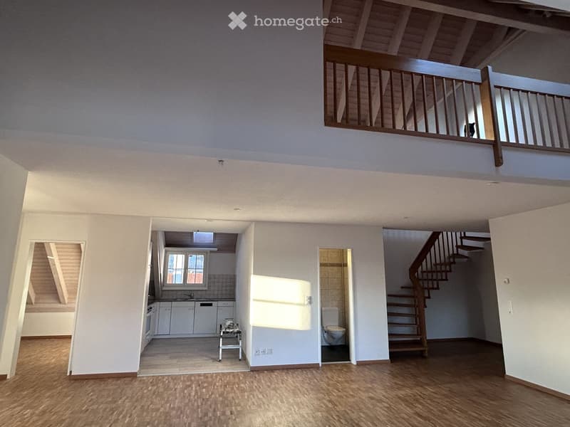 Open-plan living space with wooden flooring, high ceilings, and exposed wooden beams. Features include a staircase leading to an upper level, a kitchen area with white cabinets and appliances, a small table with a vase, and a bathroom with a toilet visibl