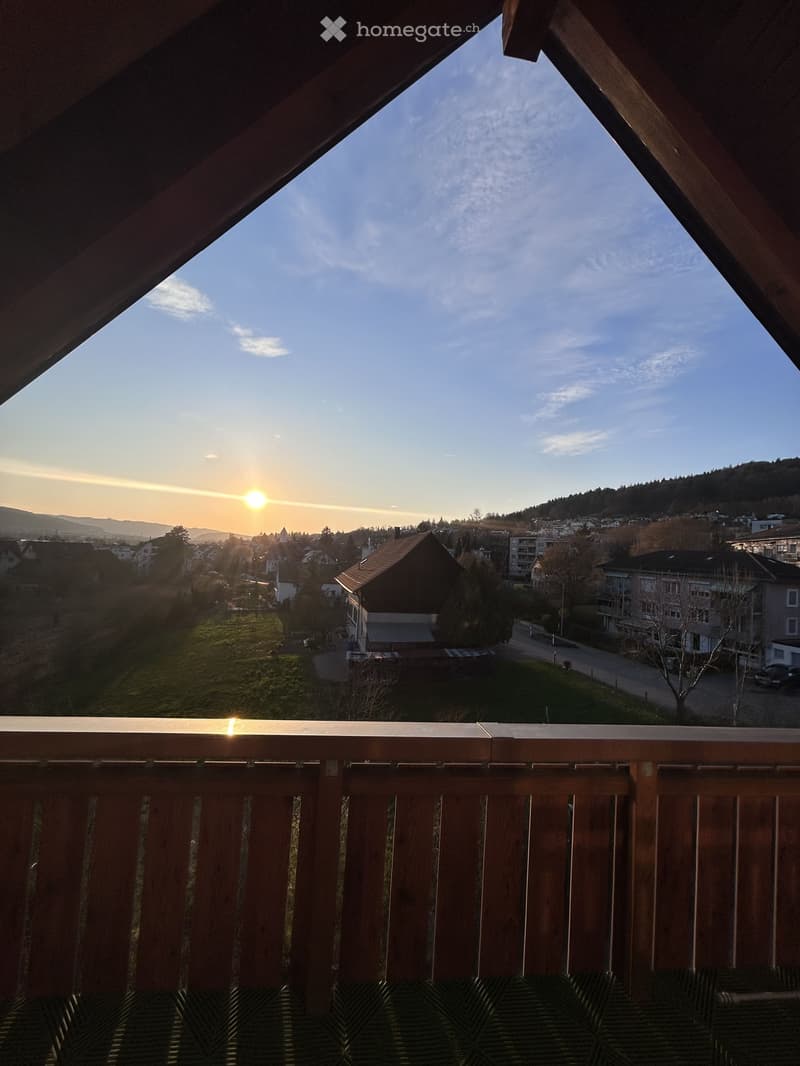 Balcony with wooden railing, overlooking a town with houses and a sunset view.
