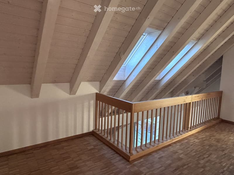 An empty attic corridor with wooden floors, white walls, and skylights above, featuring a wooden railing along the edge.