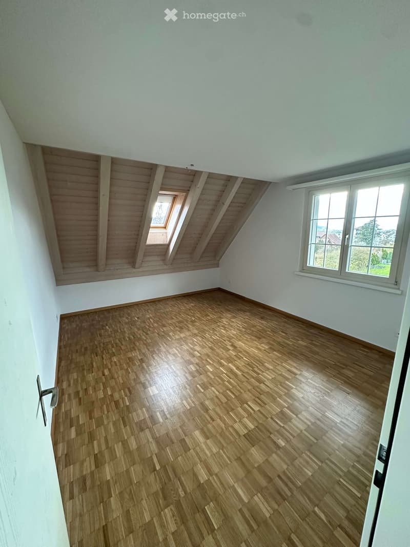 Empty room with wooden parquet flooring, white walls, and wooden ceiling beams. There is a window on the right wall and a small attic window on the ceiling.