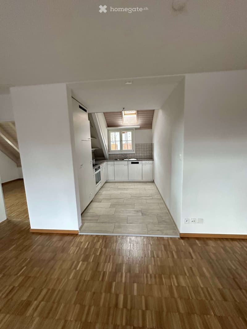 Empty room with wooden floors and white walls. There is a kitchen area with cabinets, a sink, and a dishwasher. A window and skylight are present.