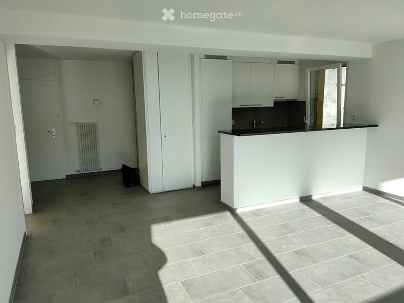 Empty room with white walls, gray tiled floor, white kitchen with black countertops, white cabinets, and a sink with a faucet.