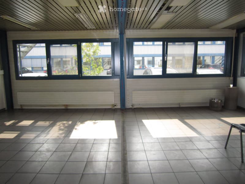 large space with windows and two pillars, tiled floor, white walls, radiators, chair