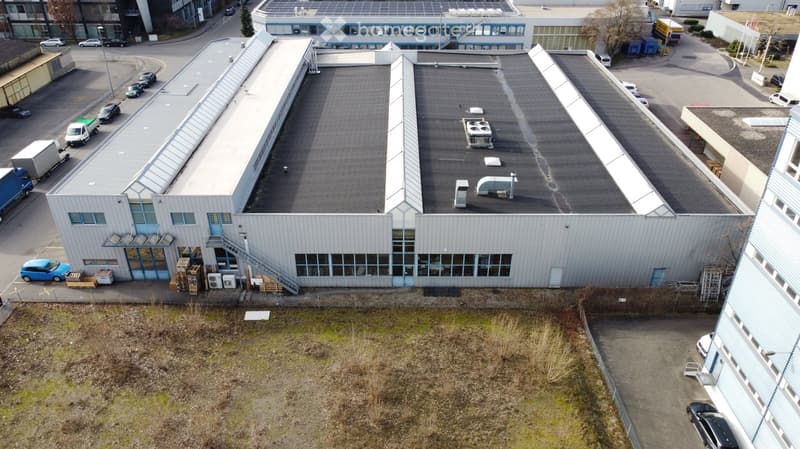 Large warehouse, glass windows, roof vents, multiple vehicles parked, concrete paved parking lot, building in the background.