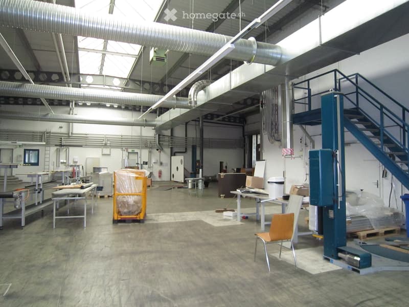 Large warehouse, concrete floor, metal shelves, equipment, office area with desks and chairs, metal staircase, pipes and ductwork on ceiling