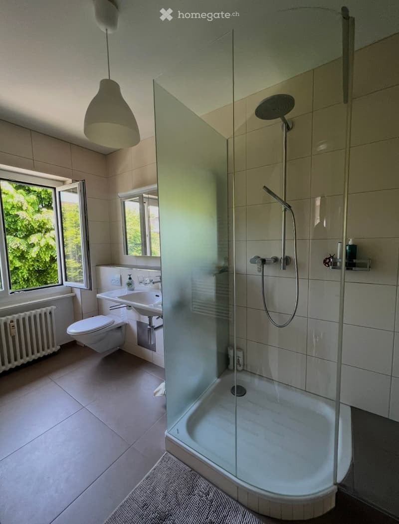Bathroom with a glass shower, white tiles, white sink, white toilet, radiator, and a window with a view of trees.