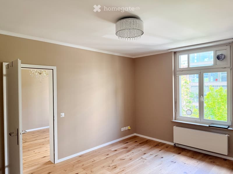 Unfurnished Room. Empty room with wooden floor, beige walls, a chandelier, and a window with a view of trees outside.