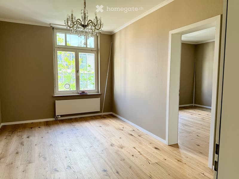 Unfurnished Room. Empty room with wooden floors, beige walls, a window with a view of trees, and a chandelier.