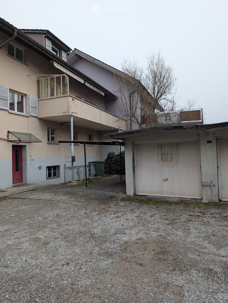 2-story residential building with balcony, red door, awning, and large parking area with covered parking and garage