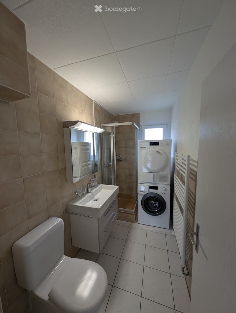 Bathroom with shower, sink, toilet, and a washing machine. White tiles and beige wall tiles.