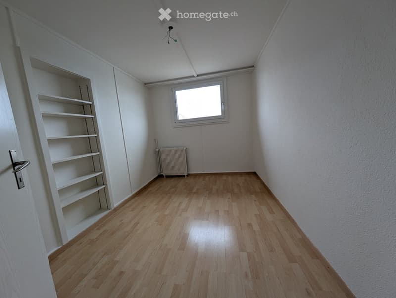 Empty room with wooden floor, white walls, white built-in shelves, small window, and white radiator.