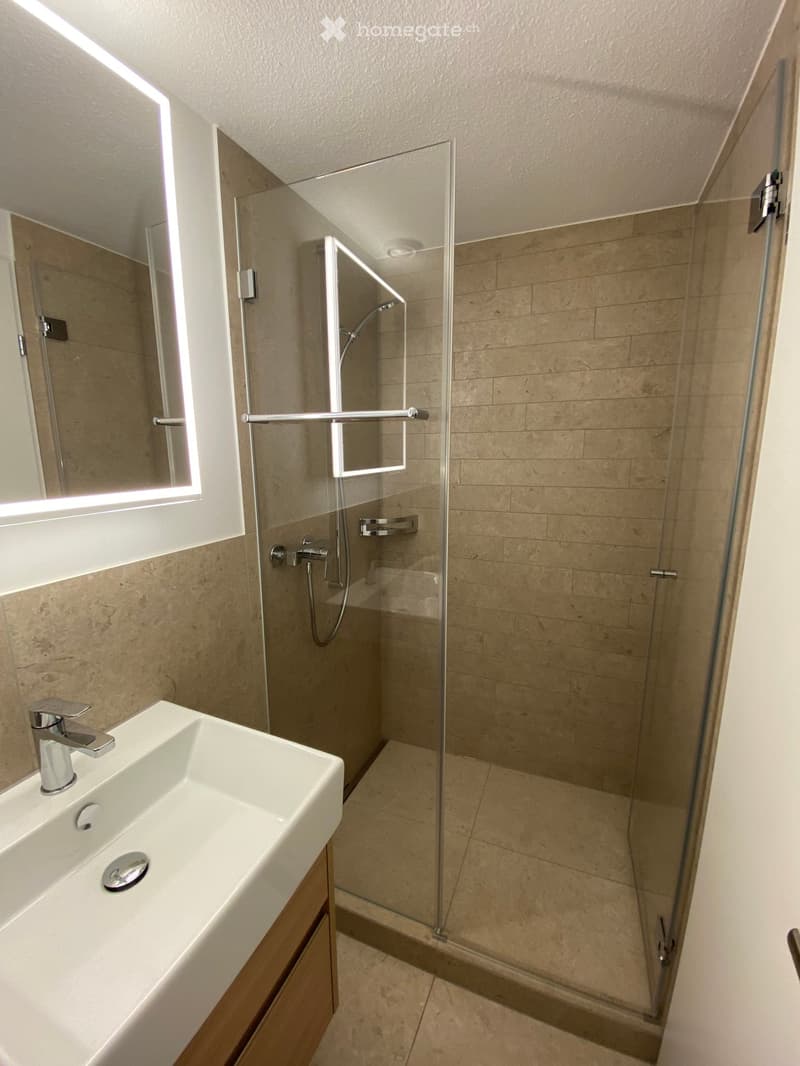 modern bathroom with a glass shower, white sink, wooden cabinet, mirror, and beige tiles