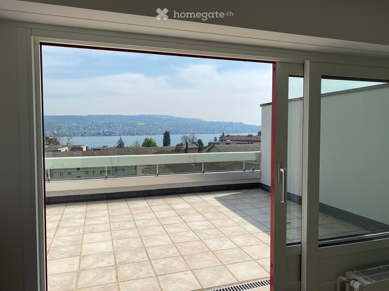 Open sliding door leading to a balcony with a tiled floor and a glass railing, overlooking a cityscape and a lake.