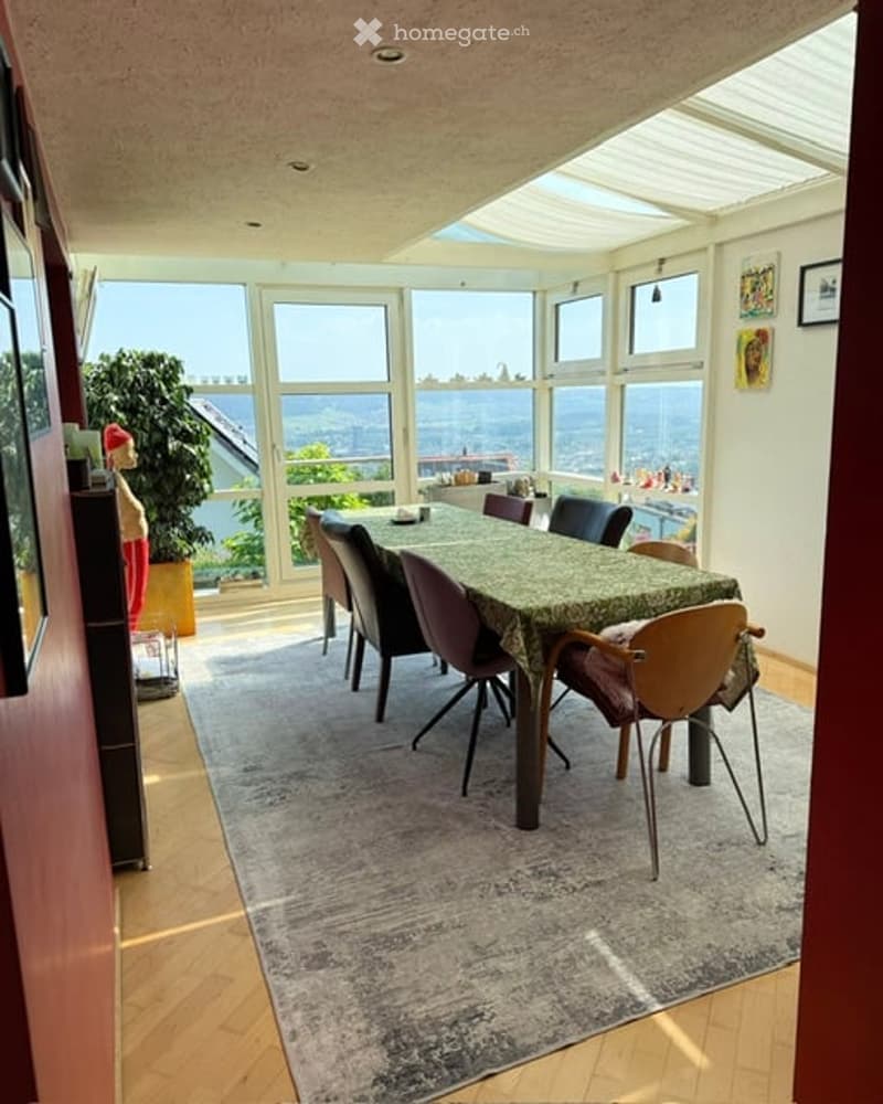 Bright dining room with large windows offering a scenic view, featuring a wooden dining table with chairs, a patterned rug, and a potted plant near the window.