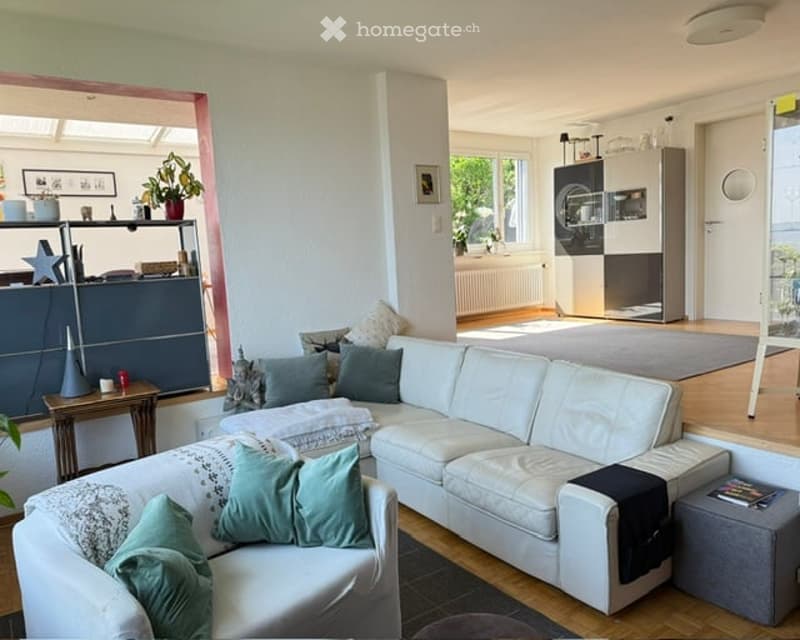Modern living room with white leather sectional sofa, multiple throw pillows, wooden coffee table, side table, bookshelf, and a small plant. The room features large windows and a neutral color palette.