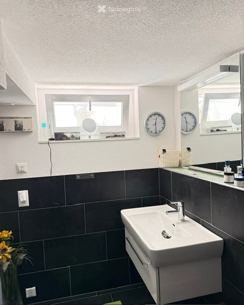Modern bathroom with dark tiled walls, white sink, large mirror, and two clocks above the sink.