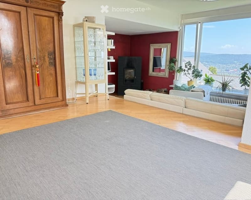 Modern living room with wooden flooring, large windows offering a scenic view, a cream-colored sectional sofa, a black fireplace, a wooden display cabinet, and potted plants on a side table.