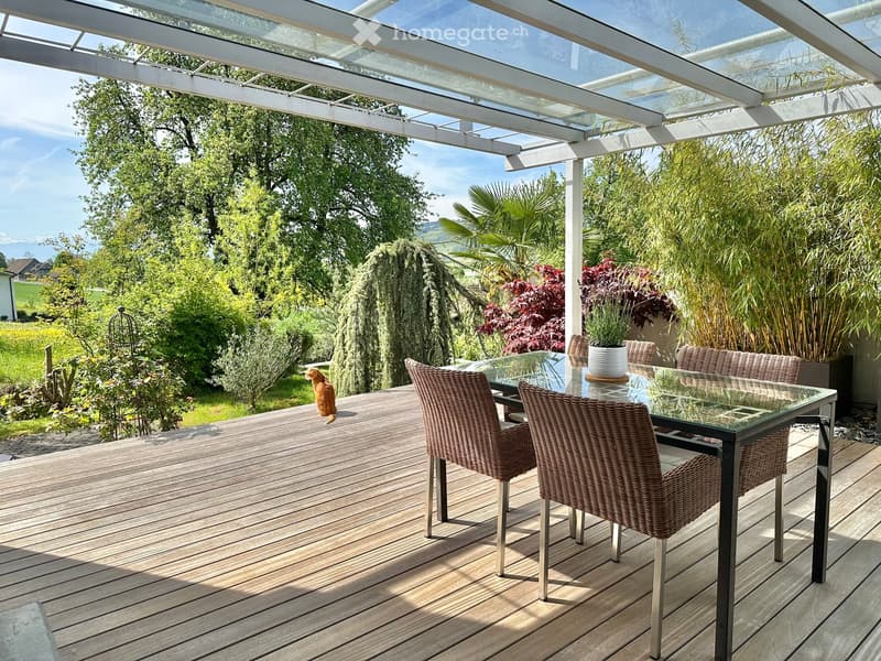 Wooden deck with a glass table and chairs, covered by a glass pergola, surrounded by plants and trees, with a view of the countryside