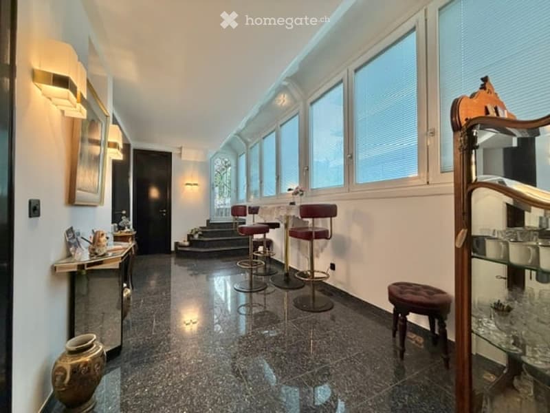 A hallway with a marble floor, white walls, and large windows with blinds. There are three red bar stools, a wooden stool, and a glass cabinet with items inside. A vase, a picture frame, and a lamp are on the left side, and a staircase is on the right sid