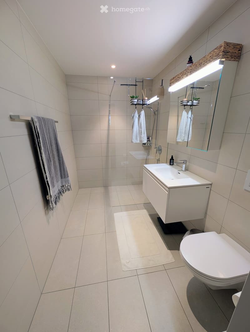 modern bathroom with white tiles, glass shower enclosure, white vanity, white toilet, towel rack, mirror with light