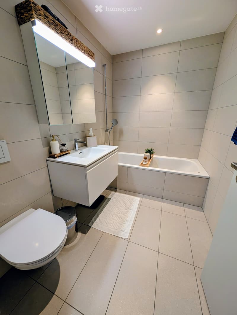modern bathroom with white tiles, white sink, bathtub, toilet, mirror with light