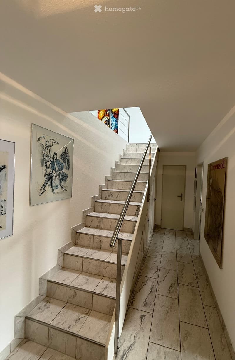 A modern staircase with marble steps and a metal railing, leading up to a landing area with a skylight and artwork on the walls.